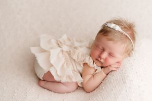 Newborn baby in cream-colored dress sleeping peacefully on white backdrop.