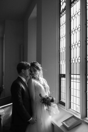 Black and white wedding photo of couple embracing by ornate church window.