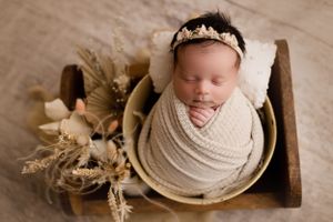 Newborn baby sleeping in wooden bowl with dried flowers and cream wrap.
