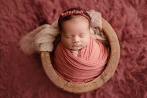 Sleeping newborn baby wrapped in pink swaddle cloth lying in wooden bowl on pink fur backdrop.