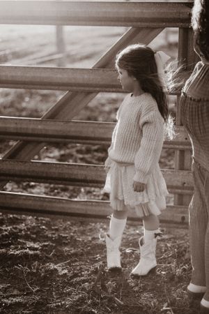 Young girl in white sweater dress stands near rustic fence in sepia tones.