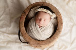 Sleeping newborn wrapped in white swaddle inside rustic wooden bowl with lace headband.