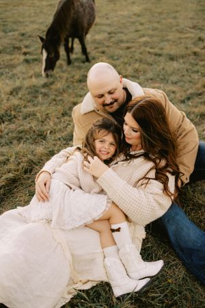A family shares a tender moment together on grassy field with a horse grazing in the background.