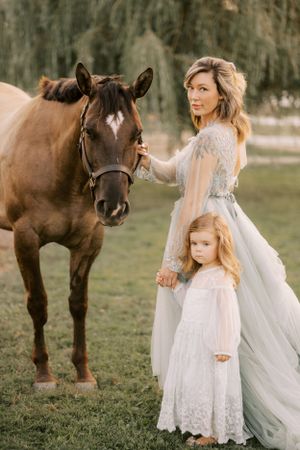 Mother in flowing dress and daughter in white lace stand with brown horse in pastoral setting at sunset.