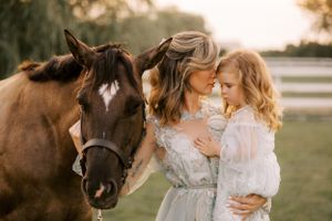 Mother holding young daughter next to gentle brown horse in dreamy outdoor setting.