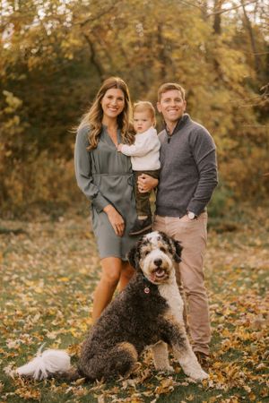 Family poses with their Bernedoodle dog among autumn leaves in a park setting during golden hour.