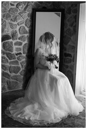 Black and white portrait of bride sitting with bouquet reflected in mirror against stone wall.