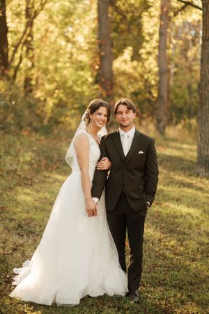 Bride and groom pose together in autumn forest wearing elegant wedding attire.