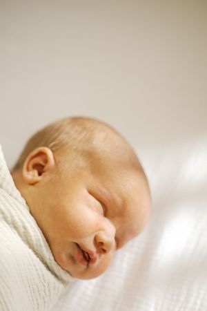 Close up of sleeping newborn baby wrapped in white blanket against light background.