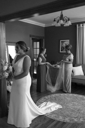 Classic black and white wedding preparation scene showing bride in white gown while others assist with final details.