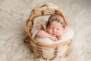 Sleeping newborn baby with floral headband nestled in woven basket on fluffy rug.