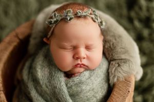 Sleeping newborn baby wrapped in gray blanket wearing delicate silver headband in wooden bowl.