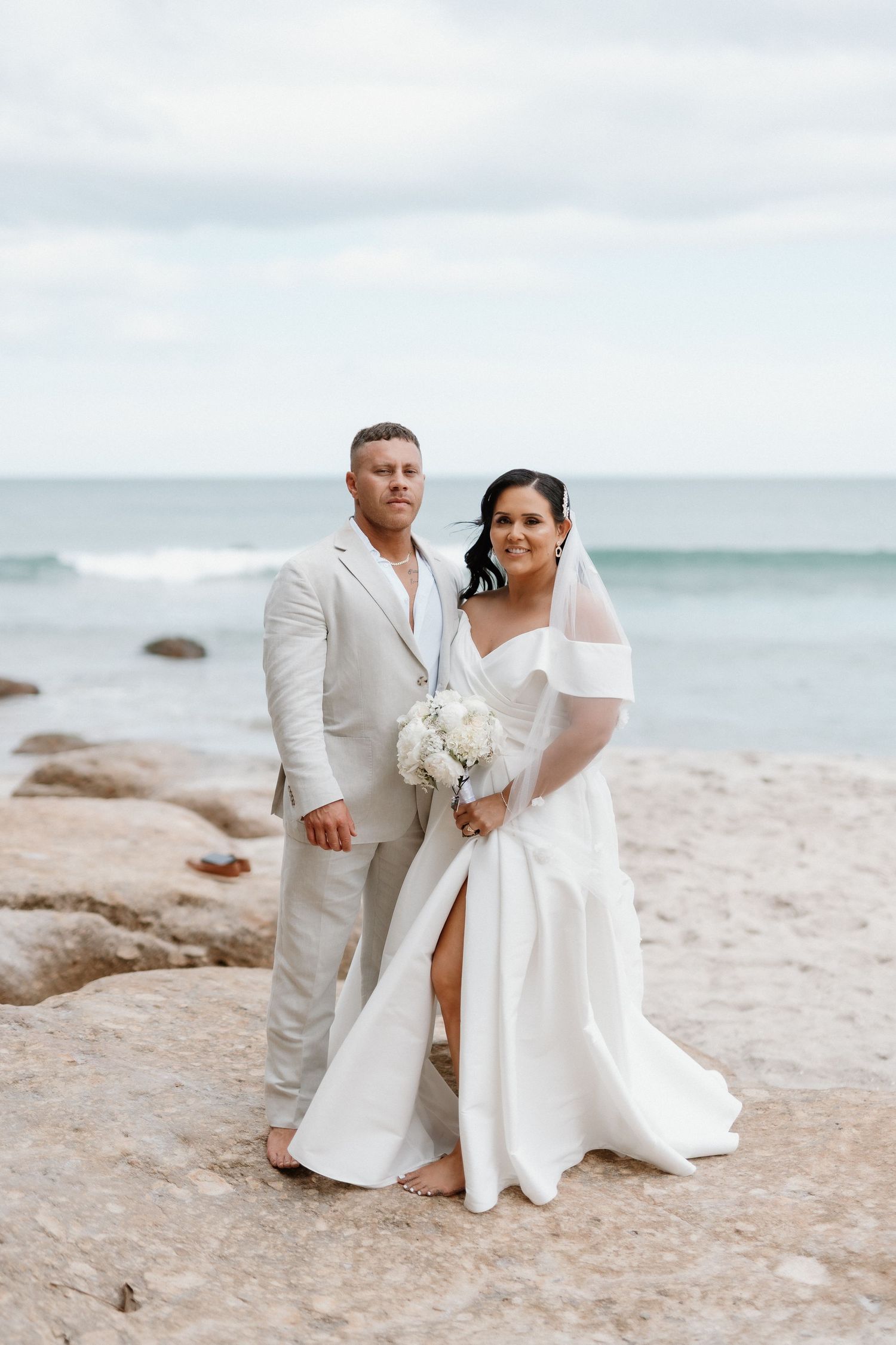 A couple in white wedding attire stands together on a rocky beach with ocean waves in the background.