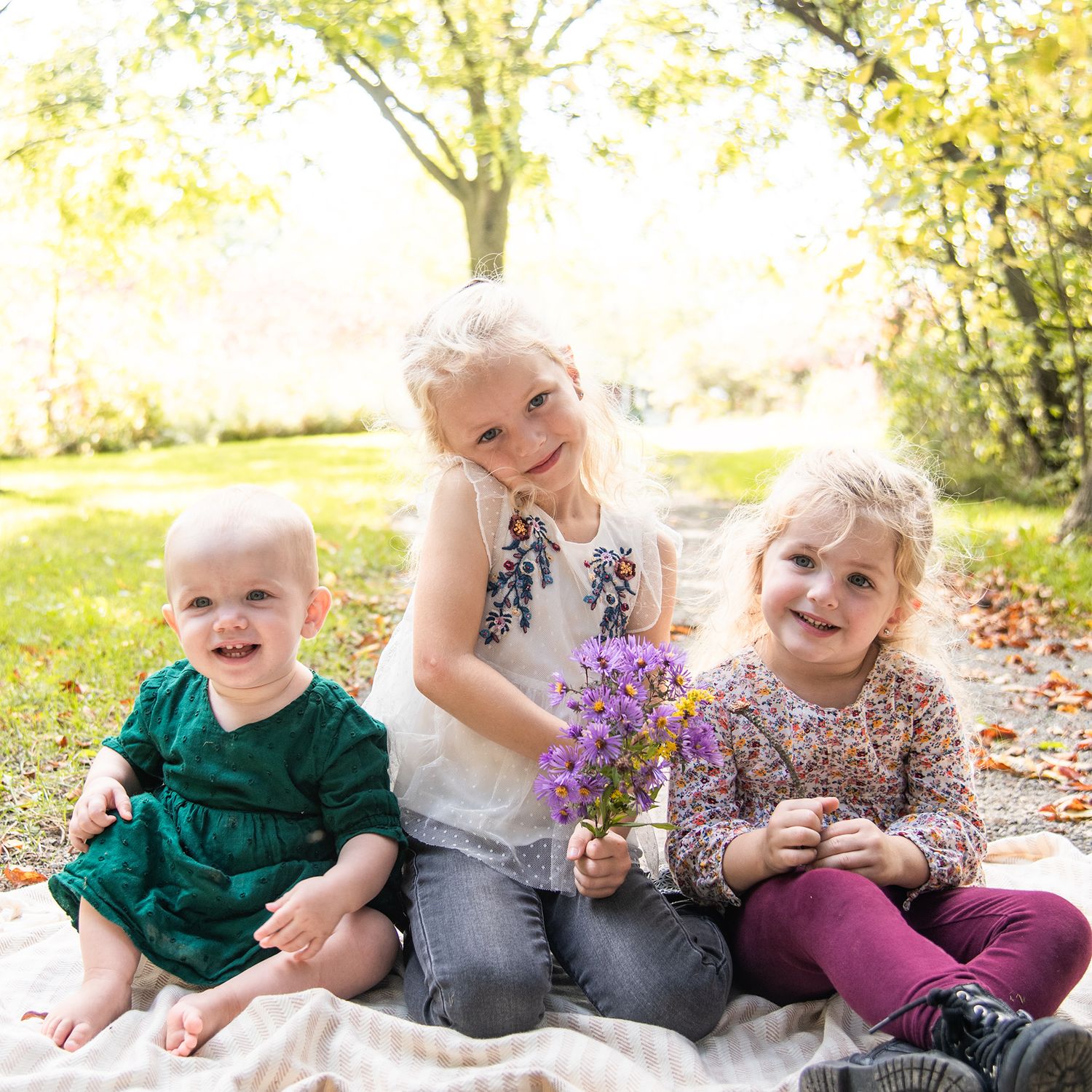 Three children sit together on a blanket outdoors in summer sunlight holding purple wildflowers.