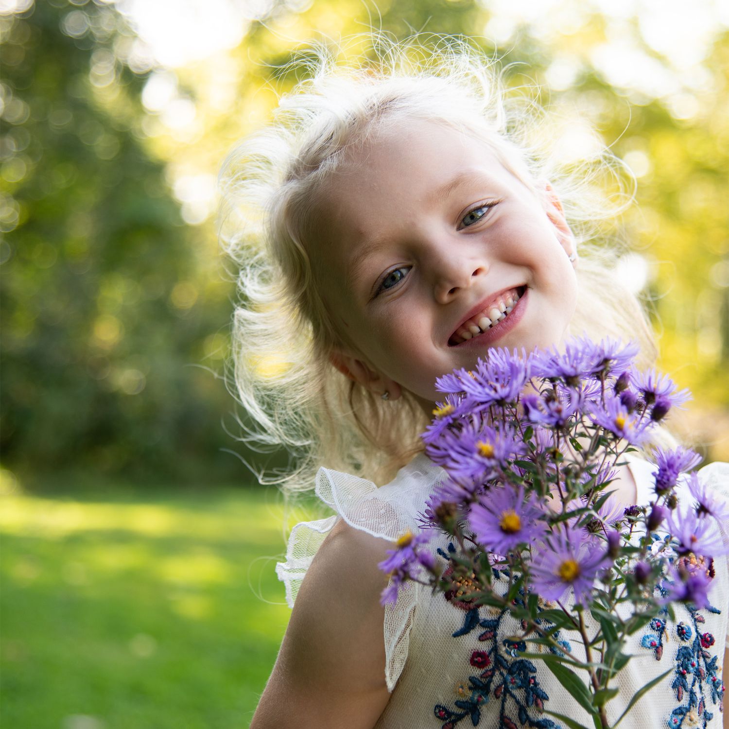 Young child holding purple wildflowers smiles brightly in sunny garden setting.