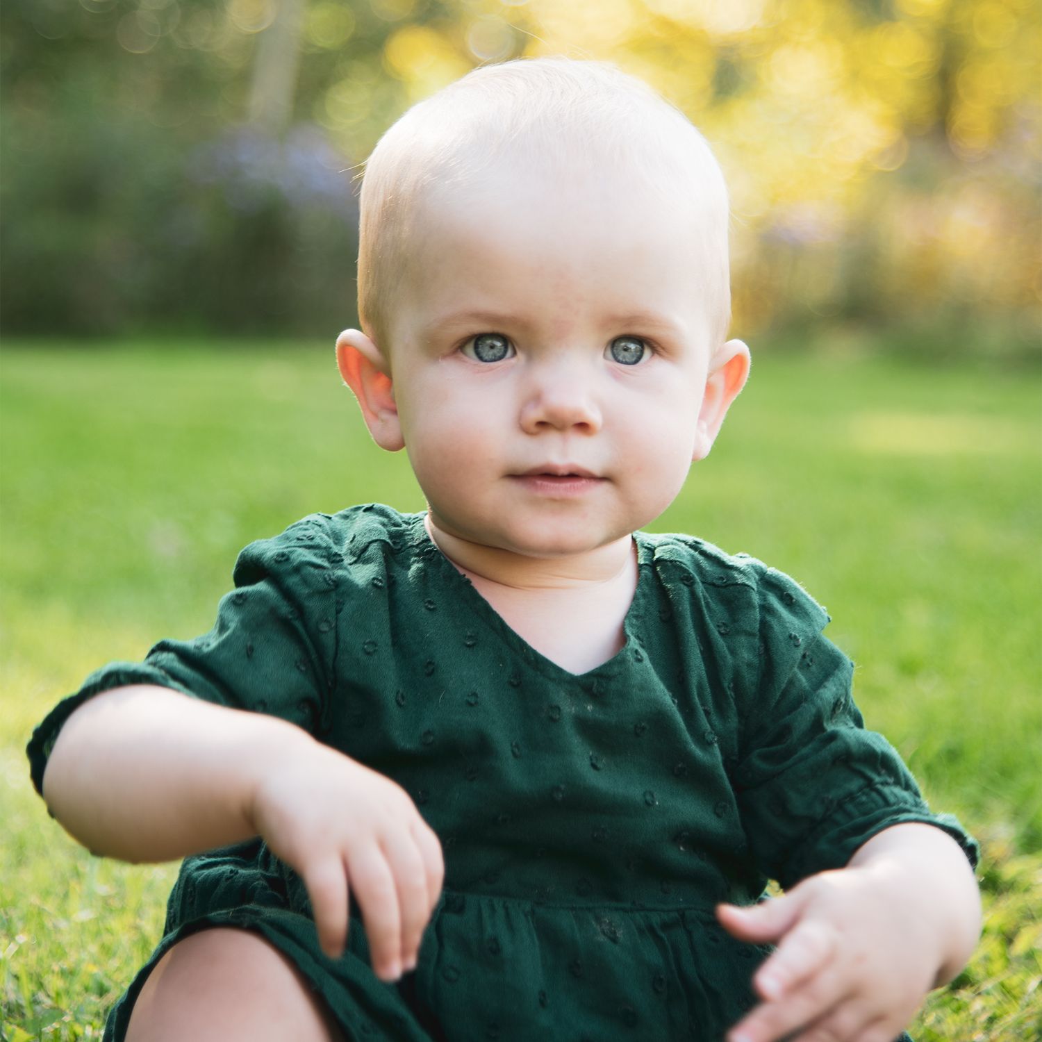 Baby in dark green outfit sitting on grass during golden hour in a garden setting.