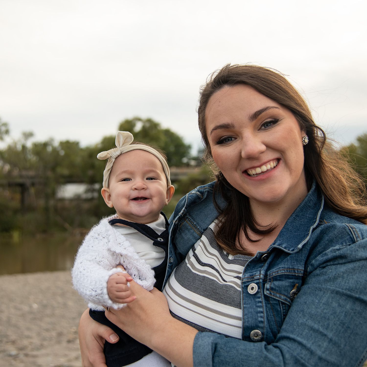 A parent and infant share a joyful moment outdoors near a lake, both wearing casual fall clothing.