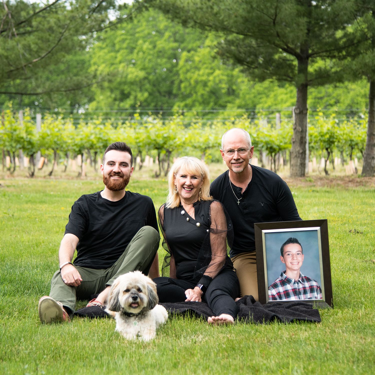 Family poses with dog and framed photo in vineyard setting.