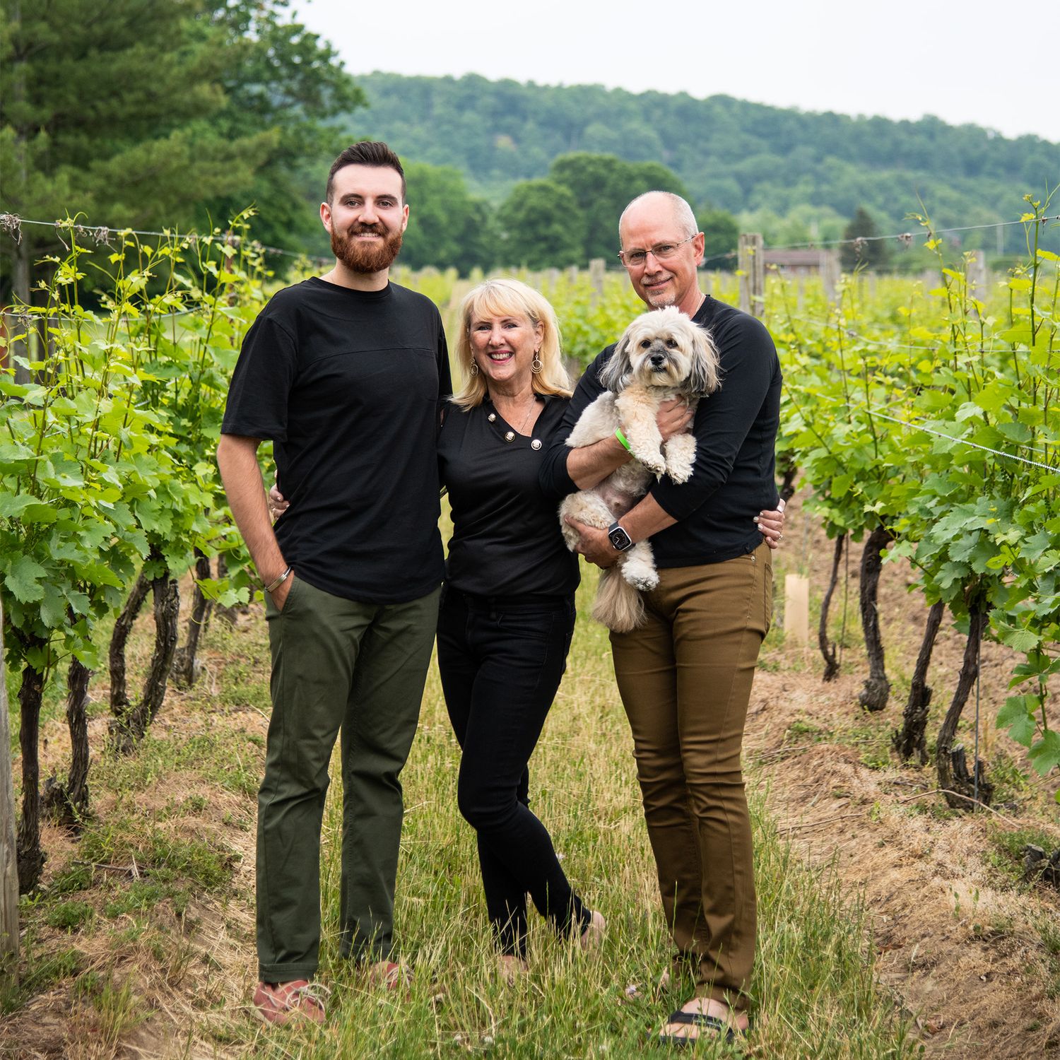 Three people and small dog pose between rows of grapevines in vineyard.