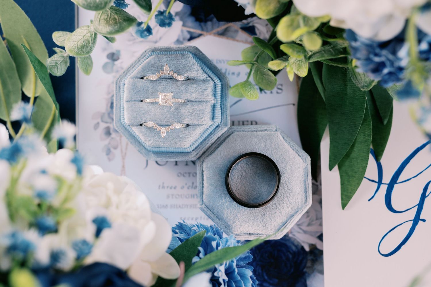 Wedding rings displayed in hexagonal boxes surrounded by blue flowers and greenery.