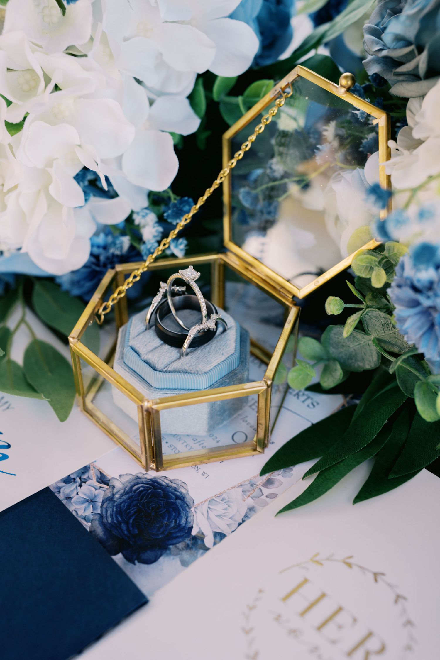 Wedding rings displayed in a hexagonal glass and gold box surrounded by white and blue flowers and greenery.