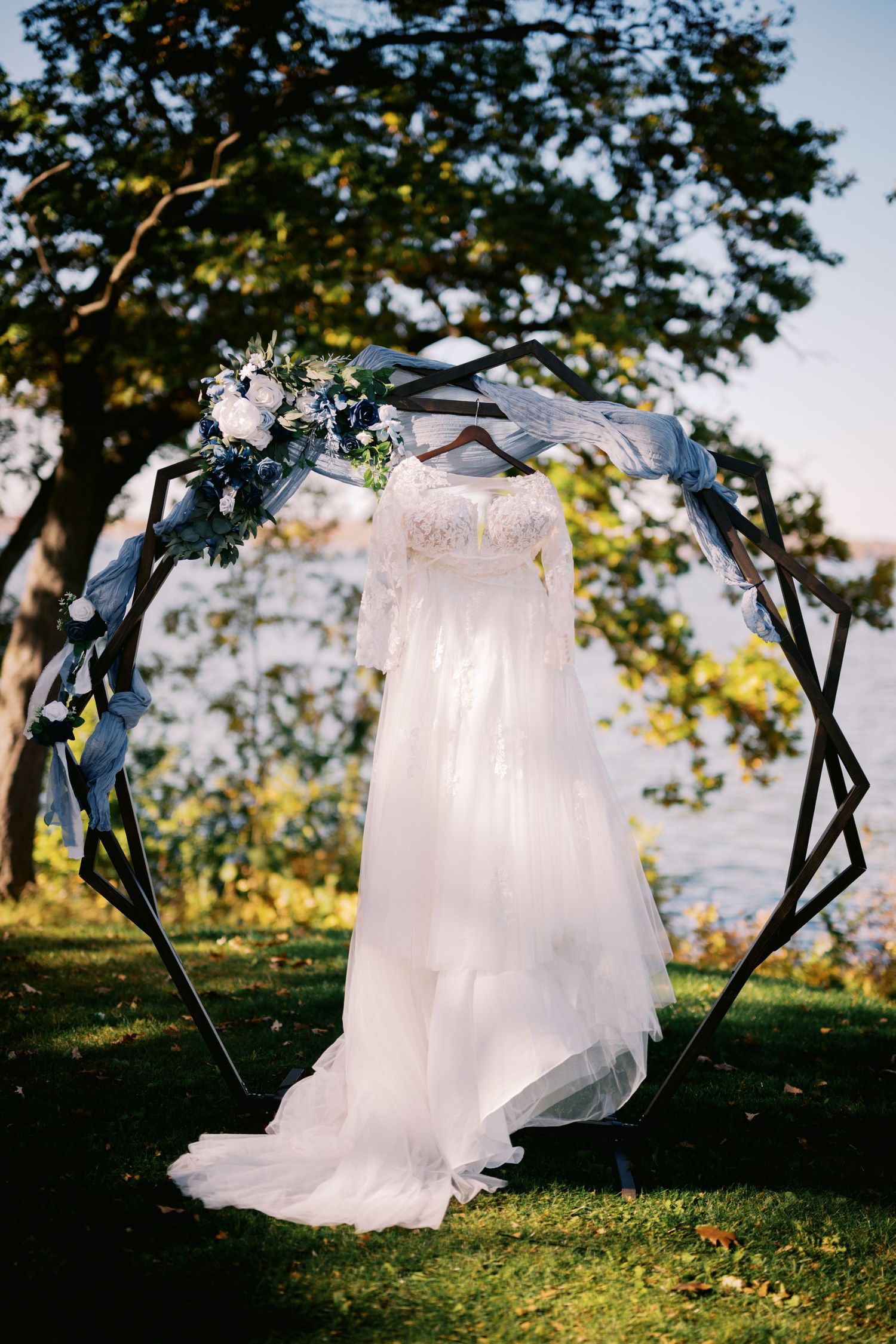 White wedding dress hanging on floral arch with blue accents against sunset sky.