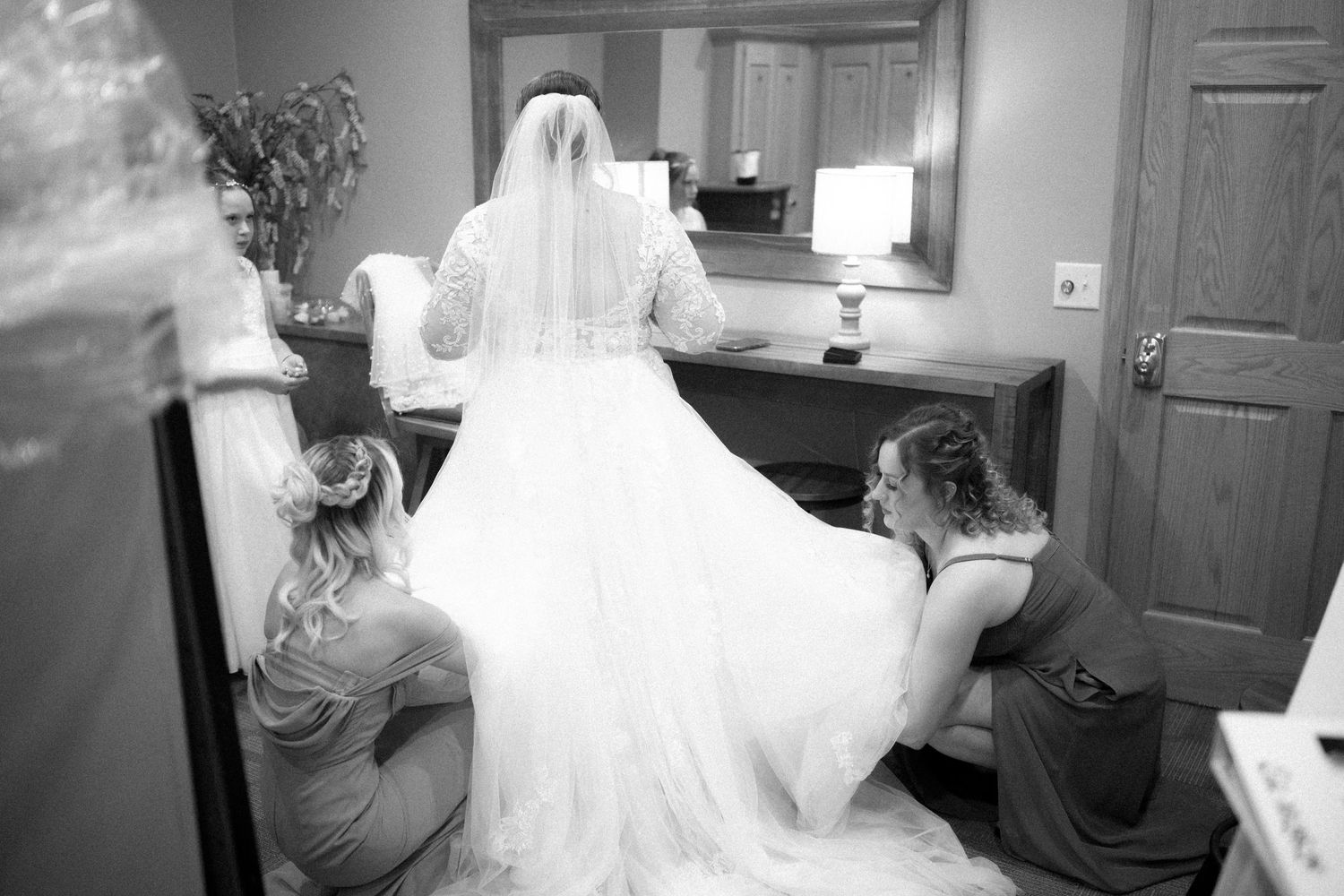 A black and white photo showing helpers adjusting a bride's wedding dress train in a dressing room.