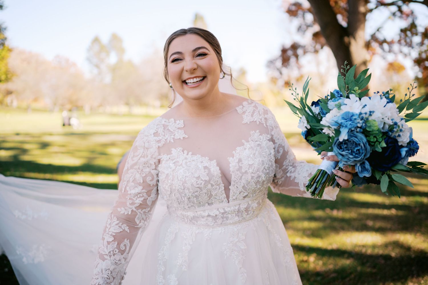 Smiling person in a lace wedding dress holding a blue floral bouquet in a sunny outdoor setting.
