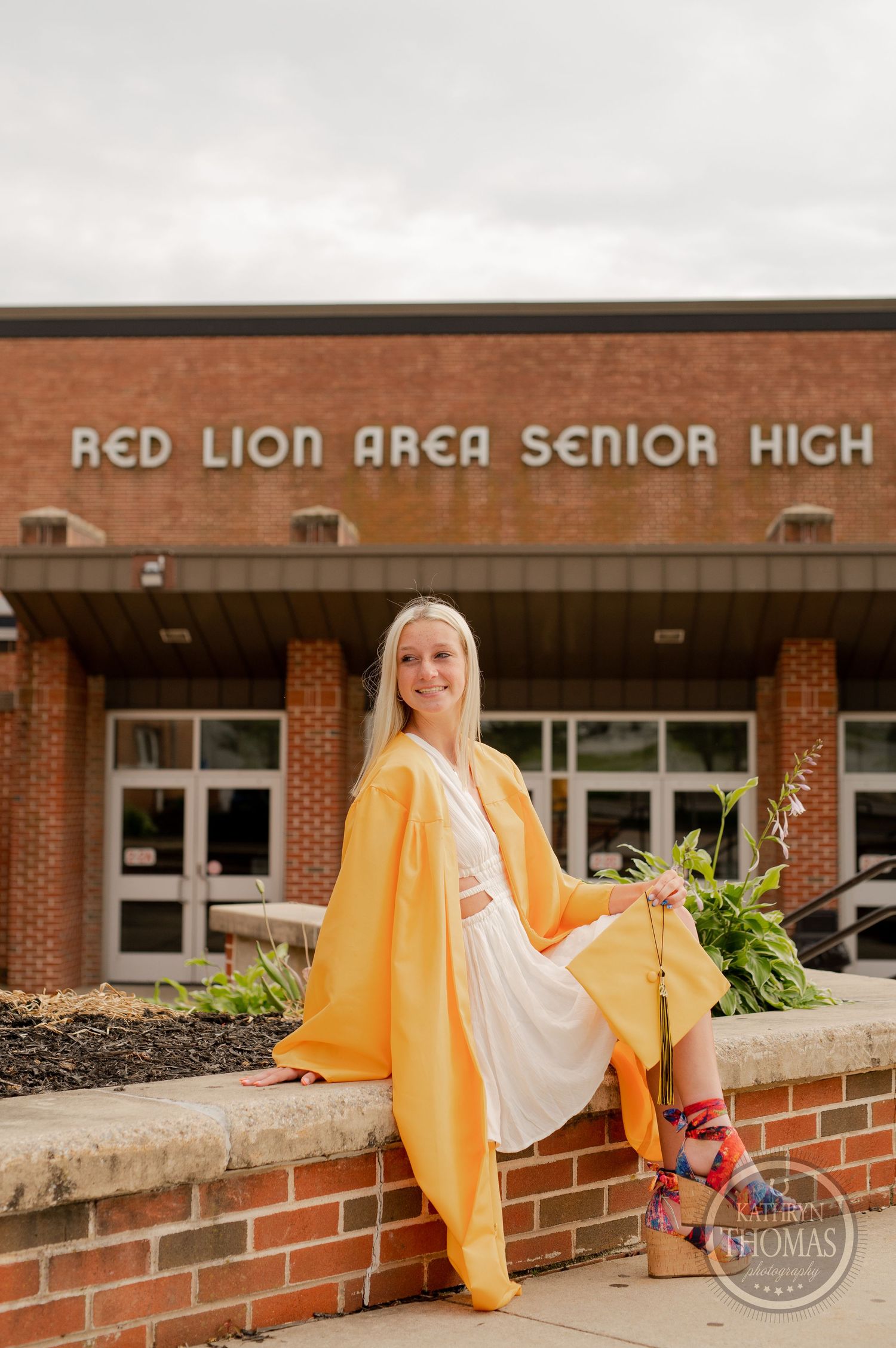 Graduate in yellow graduation gown sits gracefully on brick wall outside school entrance.