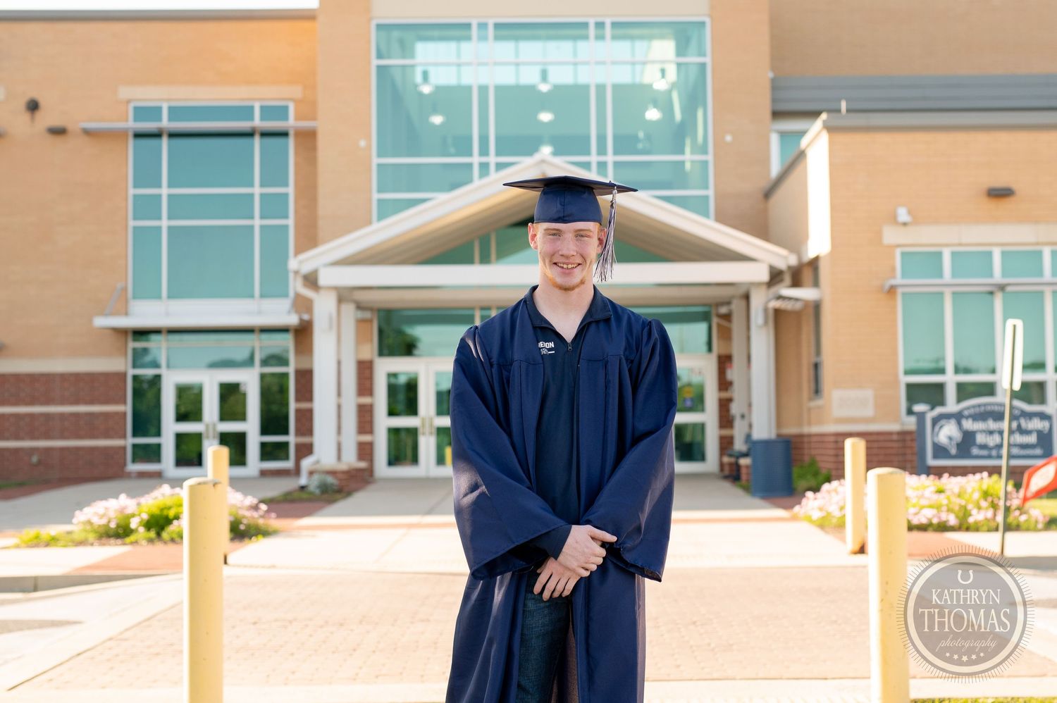 Graduate in navy blue cap and gown poses in front of modern academic building entrance.