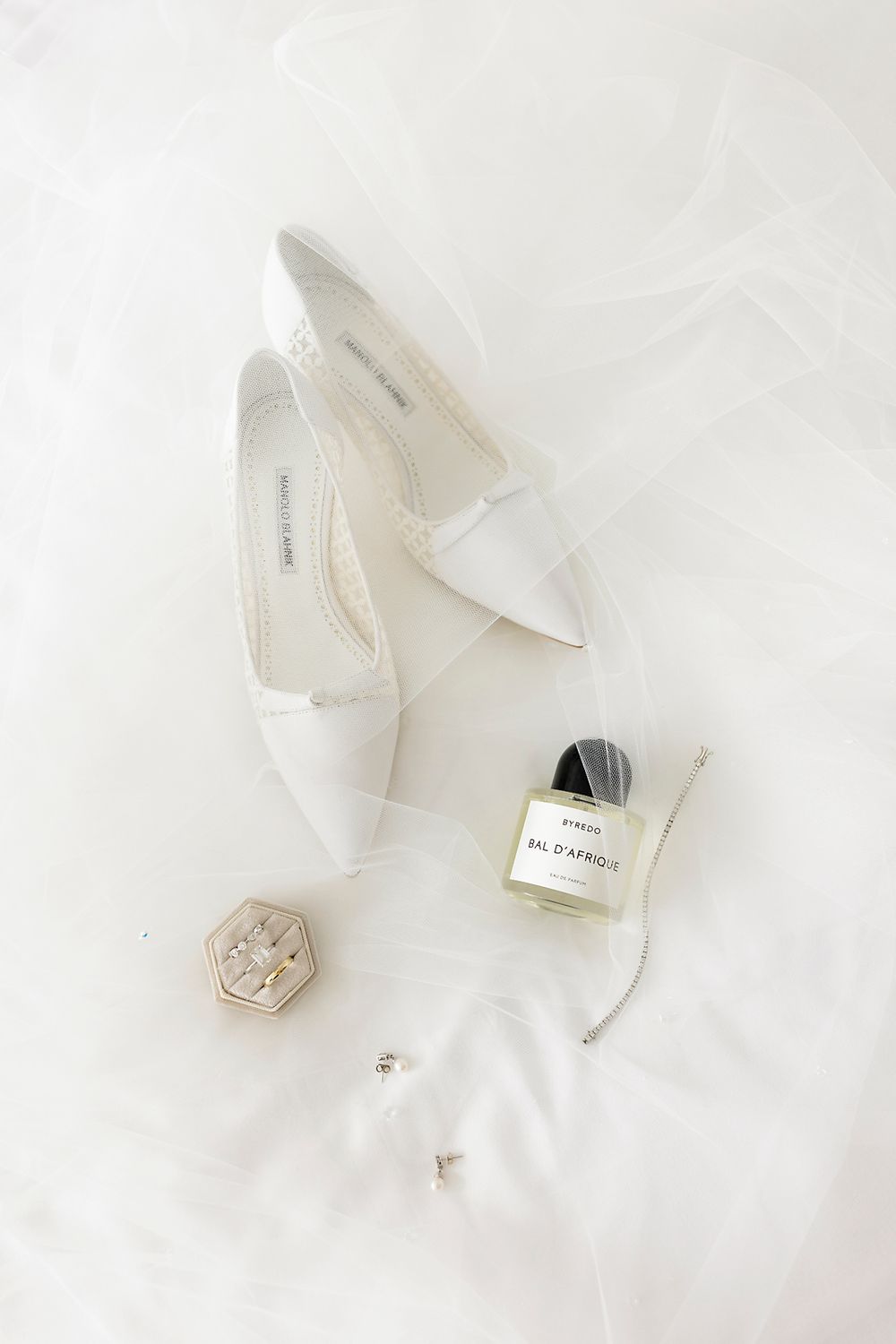 White bridal accessories including elegant pointed heels, perfume bottle, and decorative details on soft white fur background.