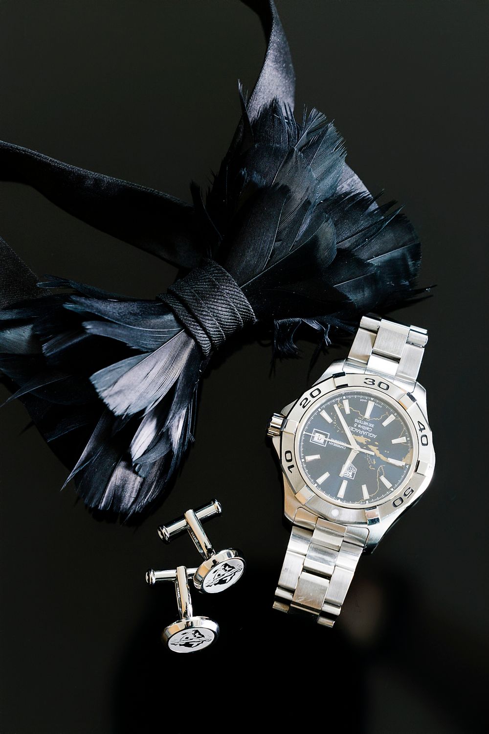 Elegant men's accessories including a silver watch, decorative cufflinks and black feather bow tie on dark background.
