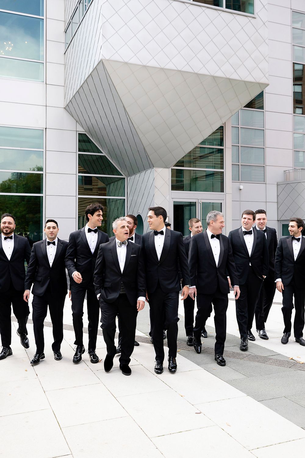 A group in formal black tuxedos and bow ties walking together outside a modern architectural building.
