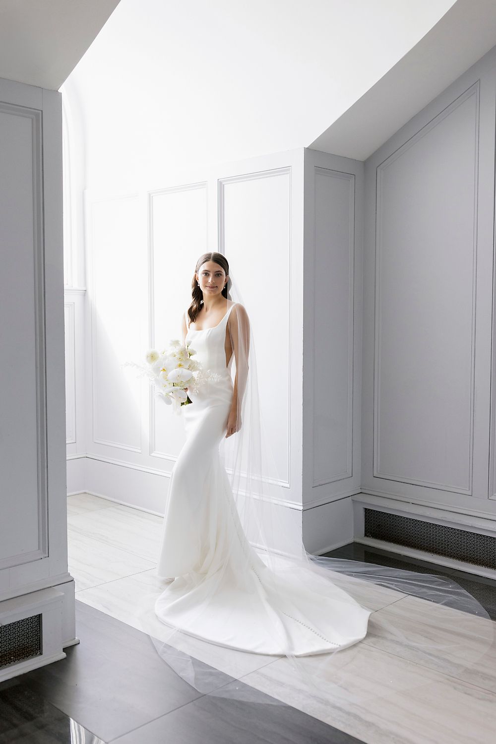A bride in an elegant white mermaid-style wedding gown stands in a bright minimalist room with white walls.