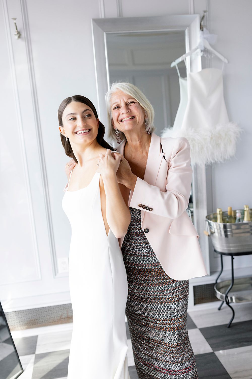 Two women share a joyful moment in a bridal boutique with a white dress hanging in the background.