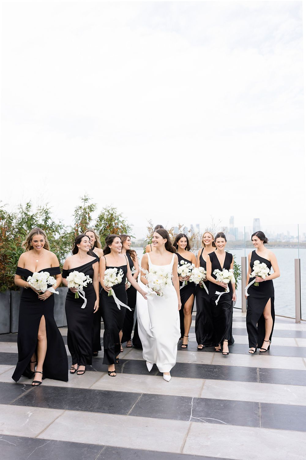 Wedding party in black bridesmaid dresses and white bridal gown walking together on a rooftop terrace with city skyline views.