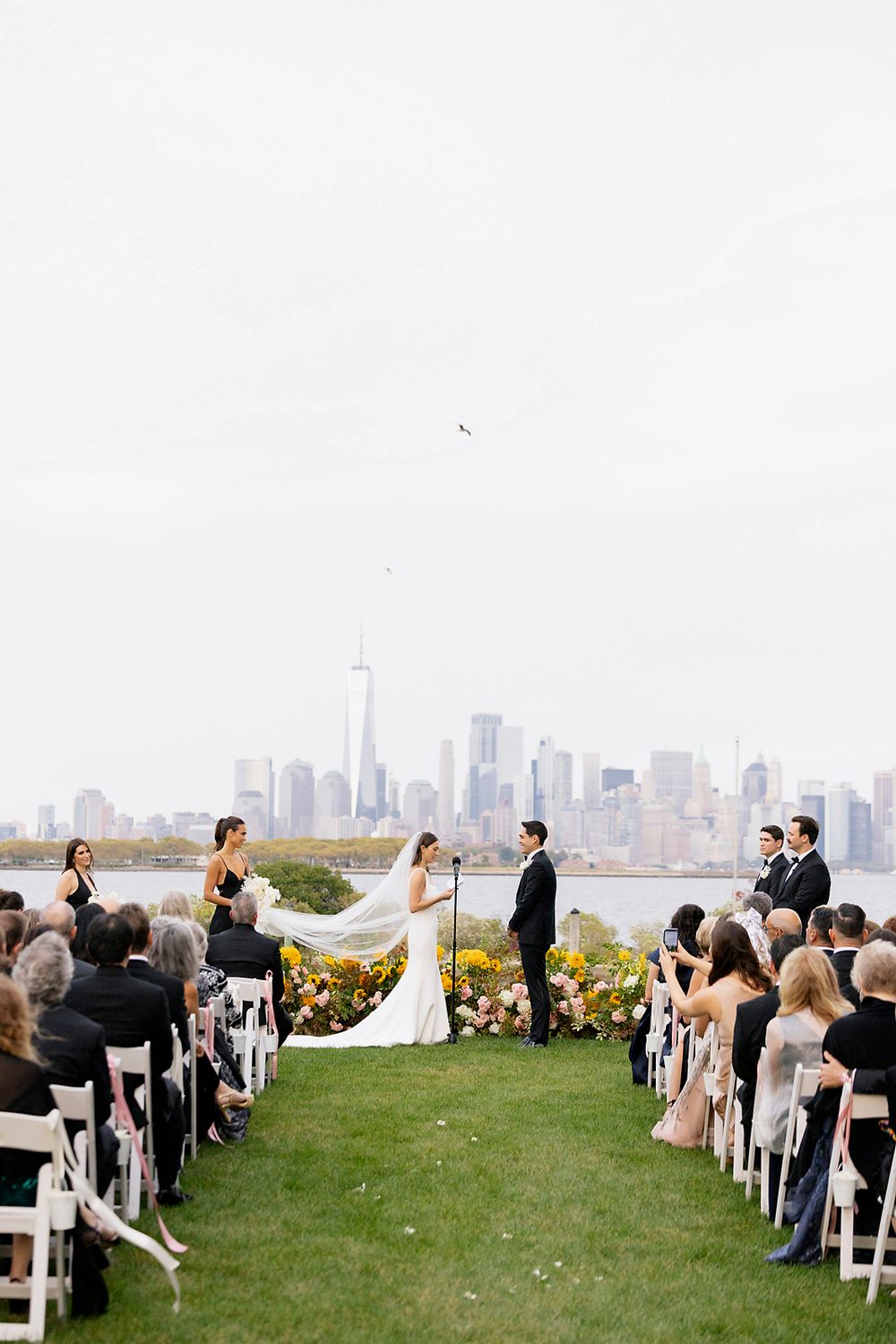 An outdoor wedding ceremony overlooking the Manhattan skyline with white chairs and yellow floral arrangements.