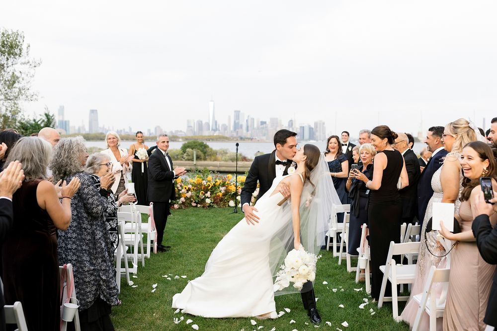 A wedding ceremony takes place outdoors with a city skyline visible in the background as guests look on.