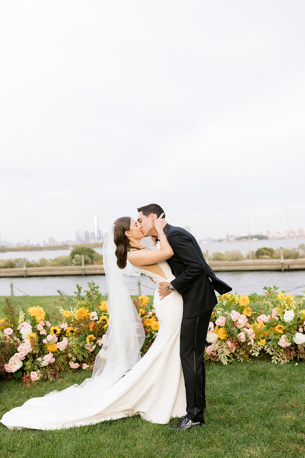 Couple shares romantic kiss on grass lawn with colorful floral arrangements and scenic waterfront backdrop.