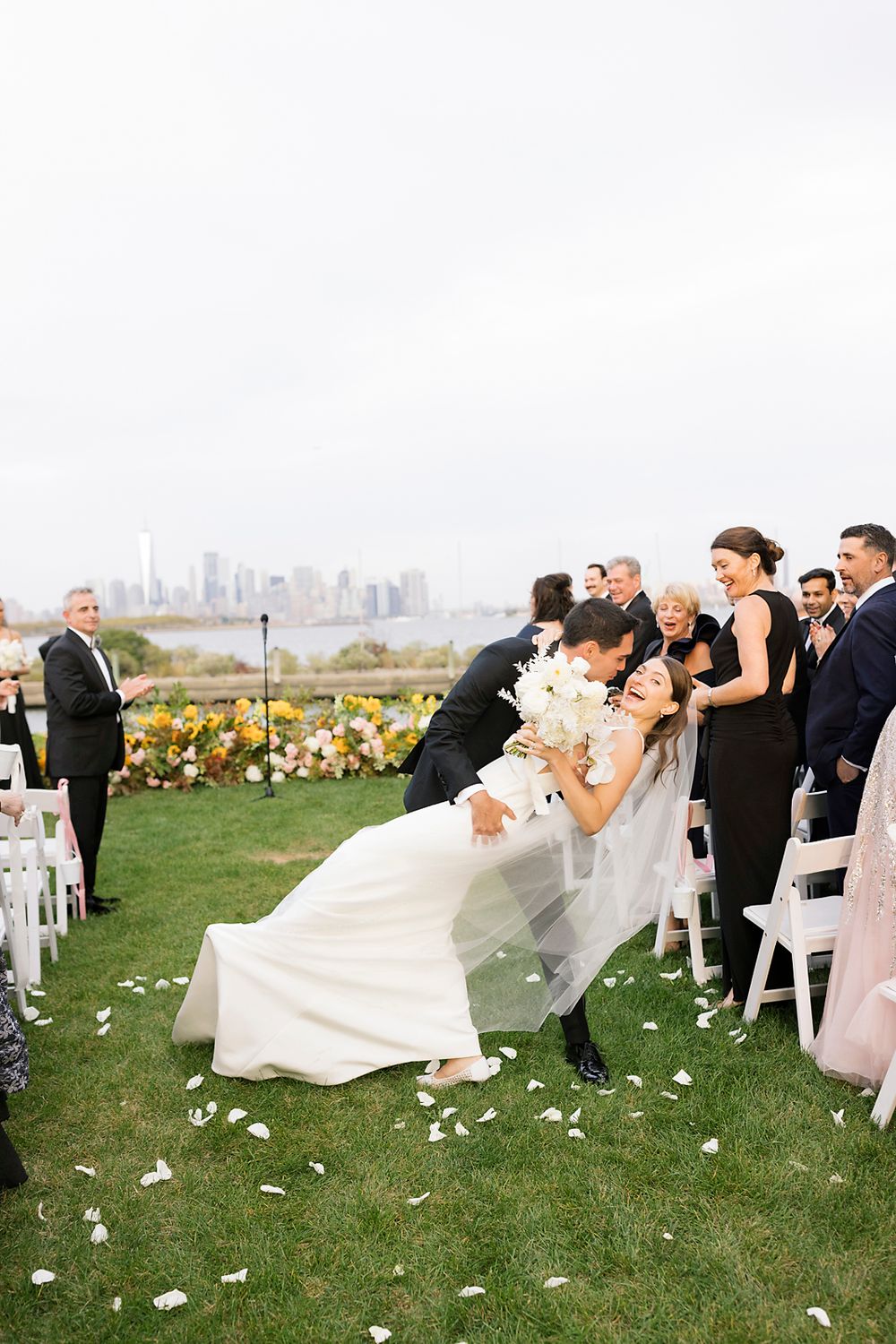 Romantic wedding scene outdoors with couple sharing a passionate kiss while guests watch on a grassy lawn with floral decor.