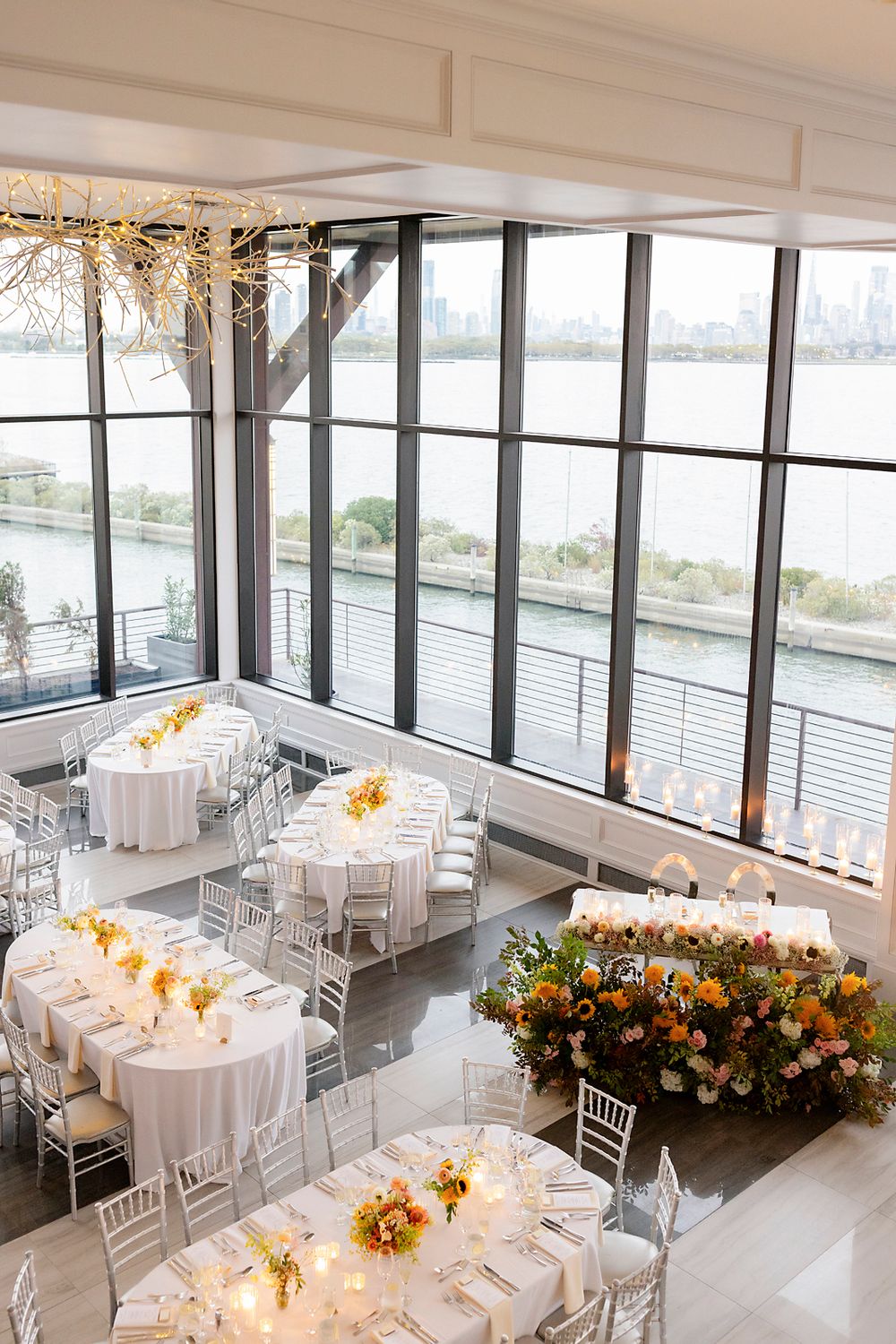 Elegant wedding reception venue with floor-to-ceiling windows, white tables, and orange floral arrangements overlooking the water.