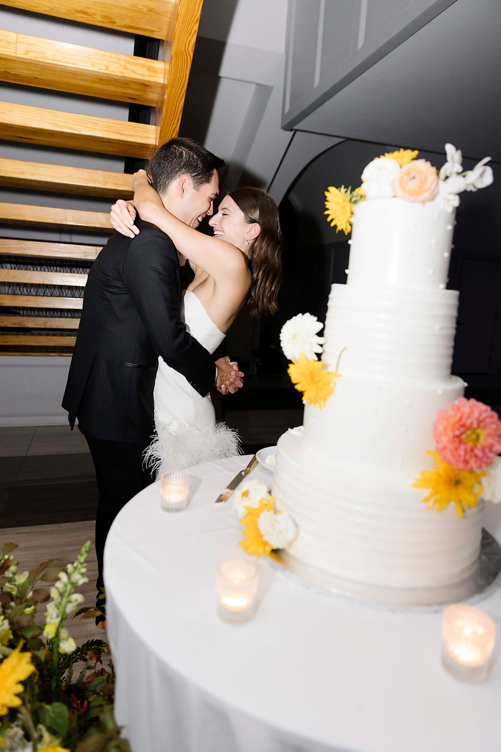 A white tiered wedding cake decorated with yellow flowers sits next to candles at a reception.
