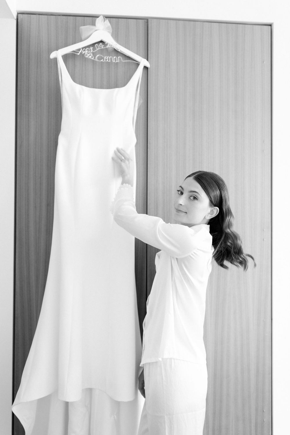 A white wedding dress hangs on display as someone in white reaches up to adjust it against a striped wall.