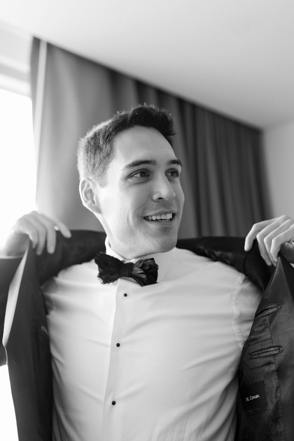 Black and white portrait of groom adjusting tuxedo bow tie and suspenders while smiling.