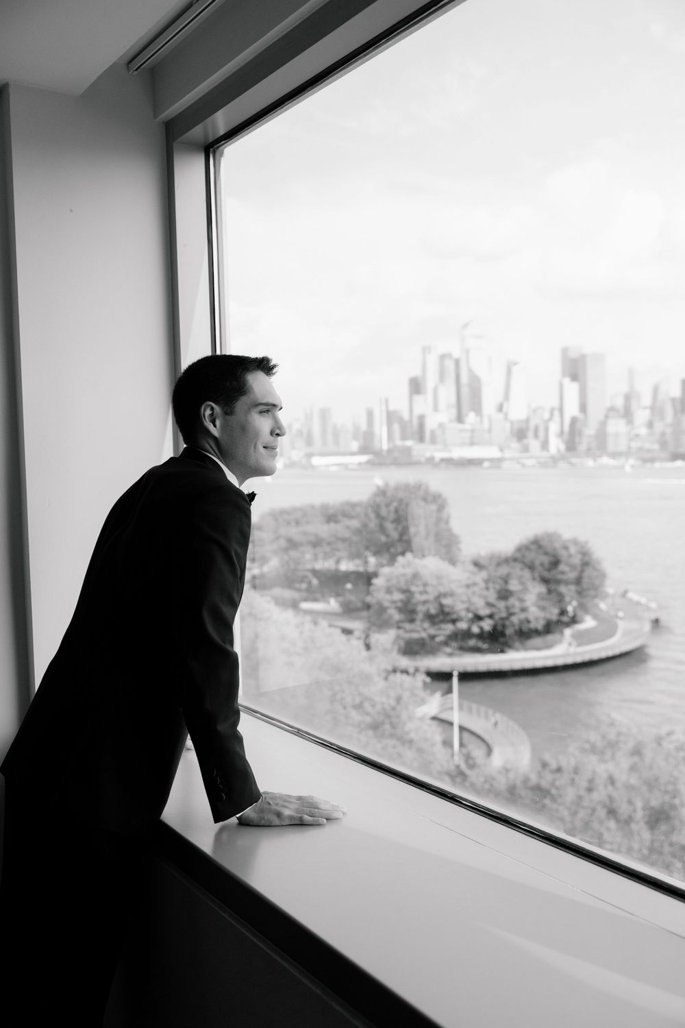 A groom in a black tuxedo looking out a high-rise window at the NYC skyline.