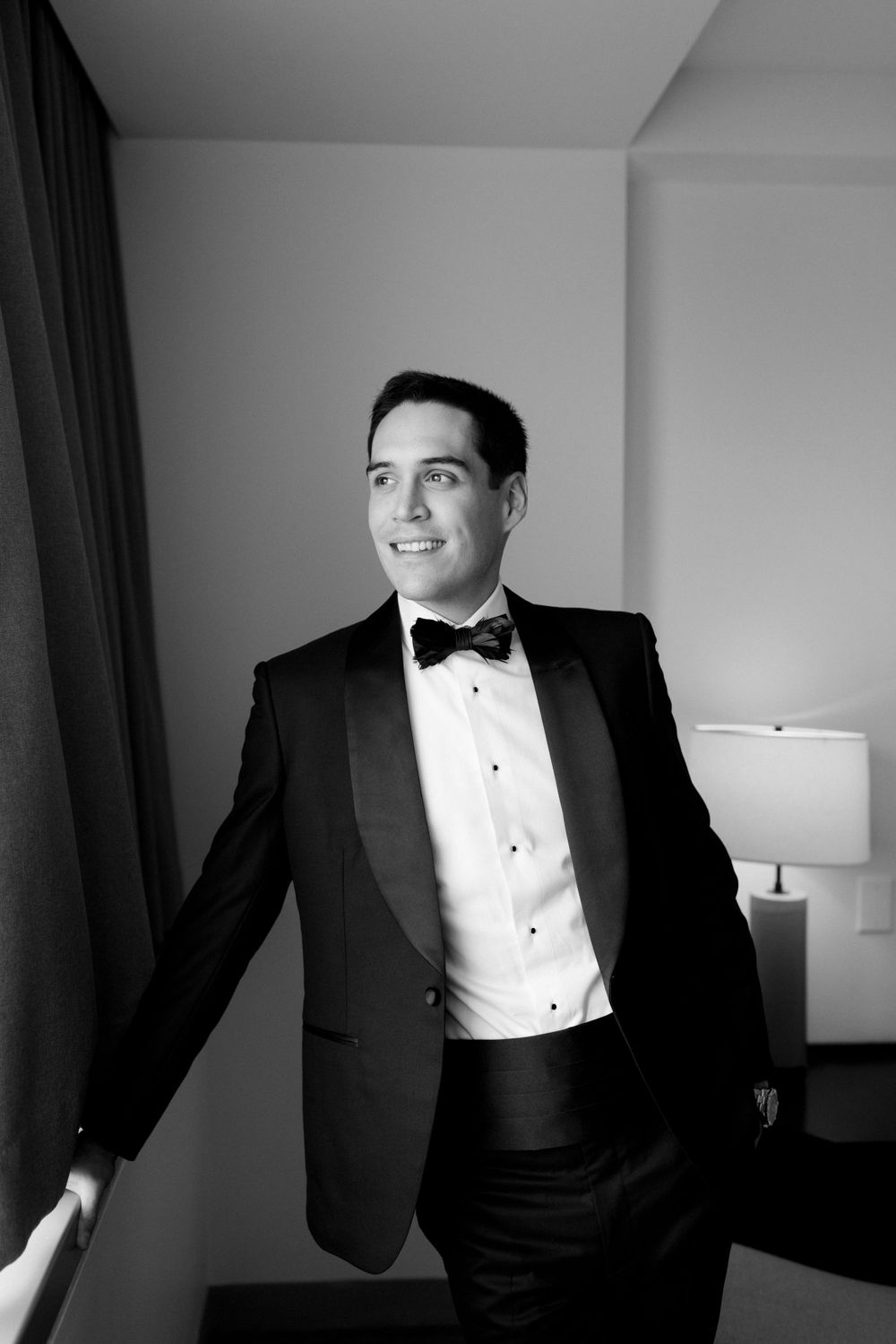 Black and white portrait of a well-dressed person in a classic tuxedo with bow tie leaning against a wall.