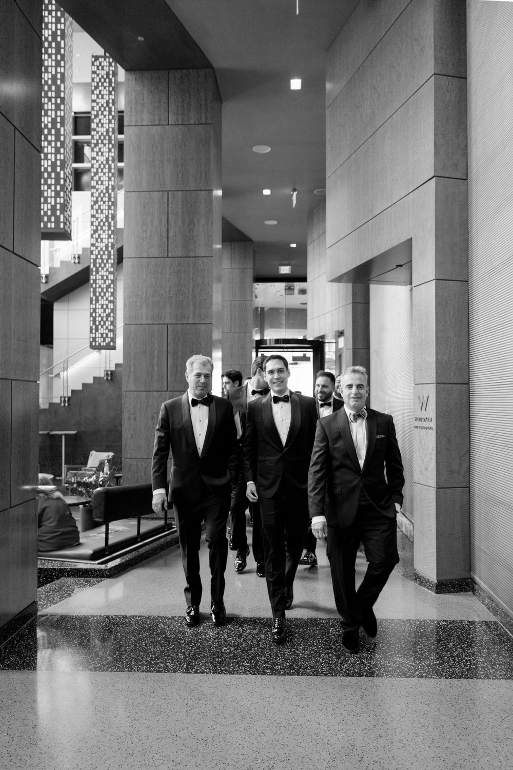 Three gentlemen in formal tuxedos walking through an elegant hallway at an event.