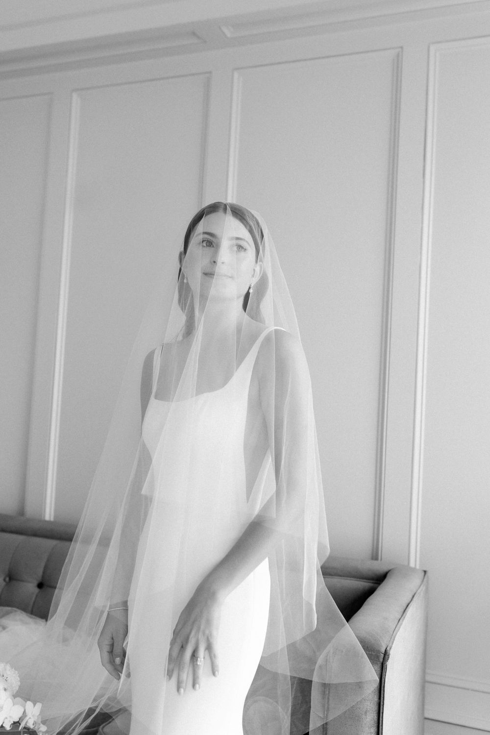 A bride in a minimalist white wedding dress stands elegantly in a black and white photograph wearing a flowing veil.