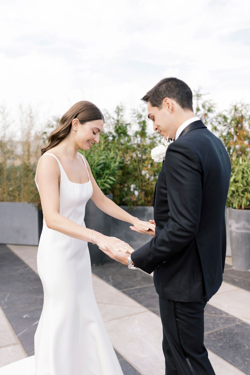 A bride in a sleek white gown and groom in classic black suit stand together during their wedding ceremony outdoors.