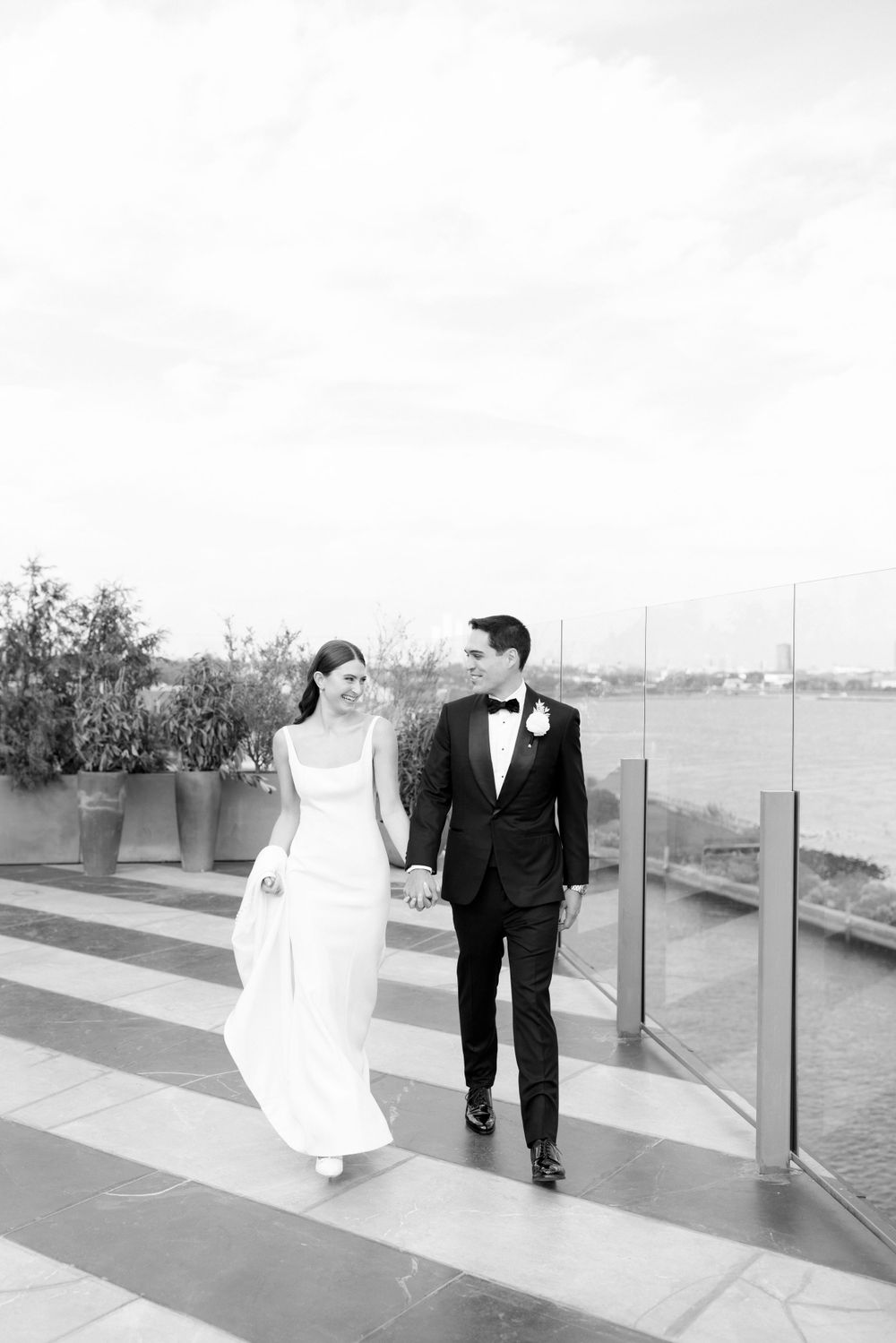 Newlyweds walking hand in hand on waterfront terrace in black and white.